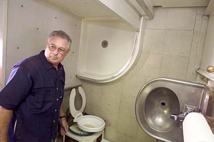 Retired Navy Capt. William Gaines standing by a toilet, with sink at ninety degrees to it - and not the ninety degrees you might be expecting - and a shower above his head. The shower tray is currently on the wall, and the walls of the shower above his head.