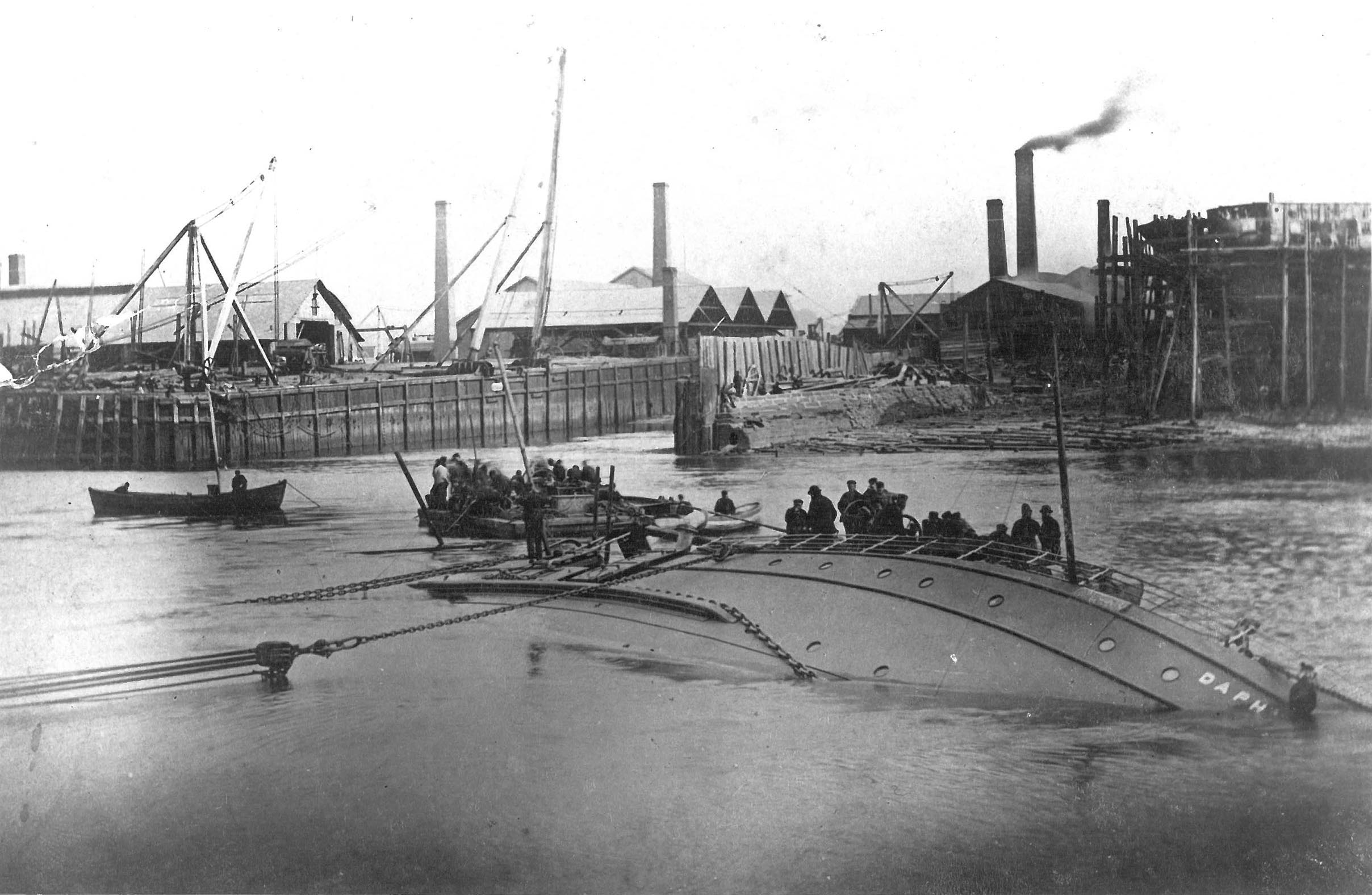 The side of a ship lies in the water with Daphne written towards the bow. Rescuers surround it. In the background is a shipyard