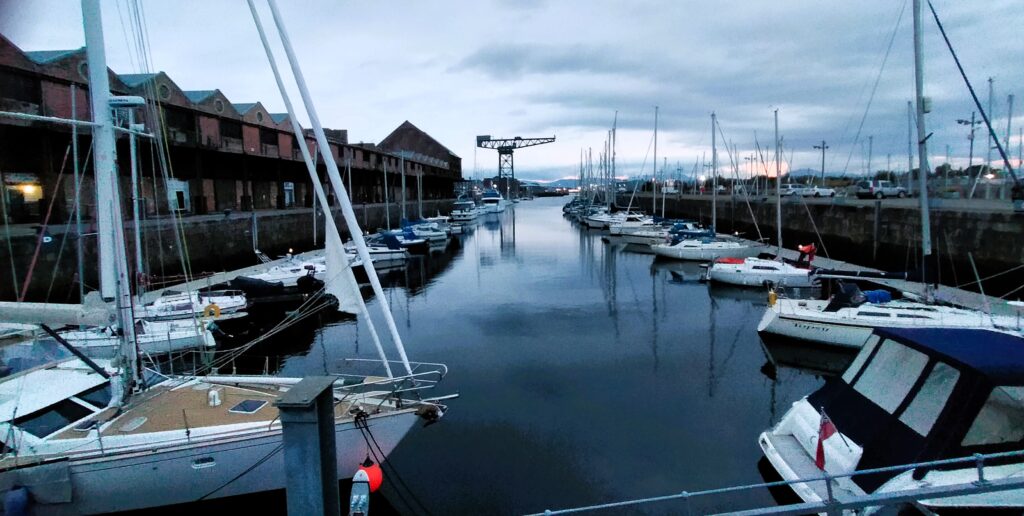 James Watt dock in Greenock. Warehouses line the left hand side and a hammerhead crane is visible in the background. Yachts line the dock against pontoons.