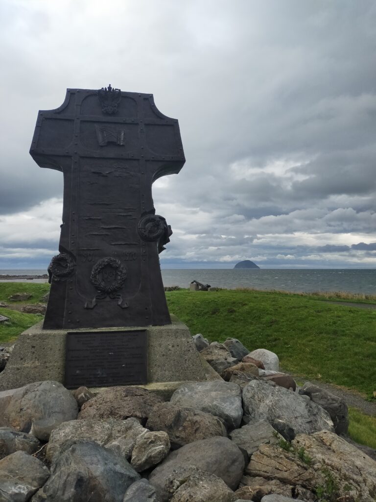 A cross with several carvings on it stands by the shore, overlooking the conical lump of Ailsa Craig